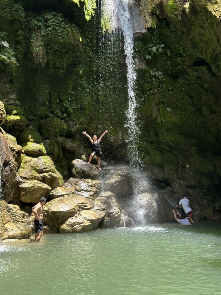Pam at a waterfall with arms raised, friends swimming below